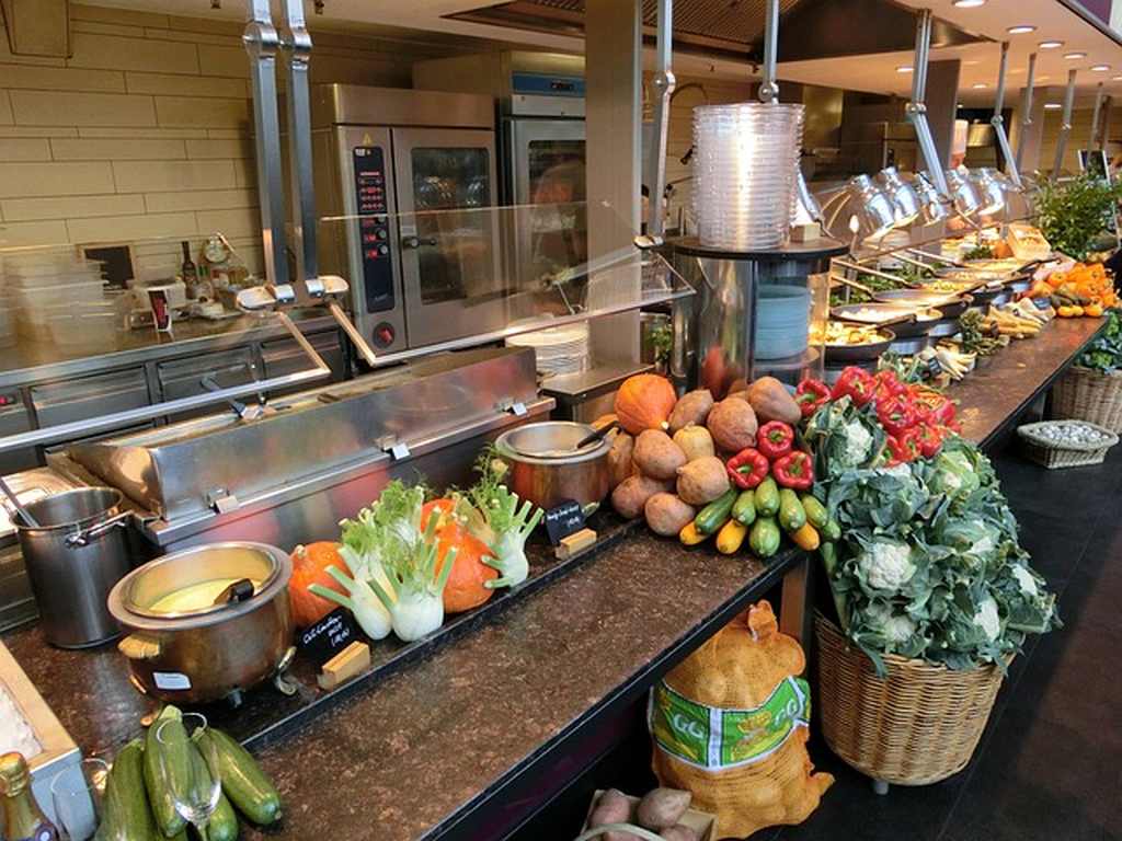 Balcão com muitos legumes e verduras, e grandes panelas de alimento. Atrás do balcão está uma cozinha industrial