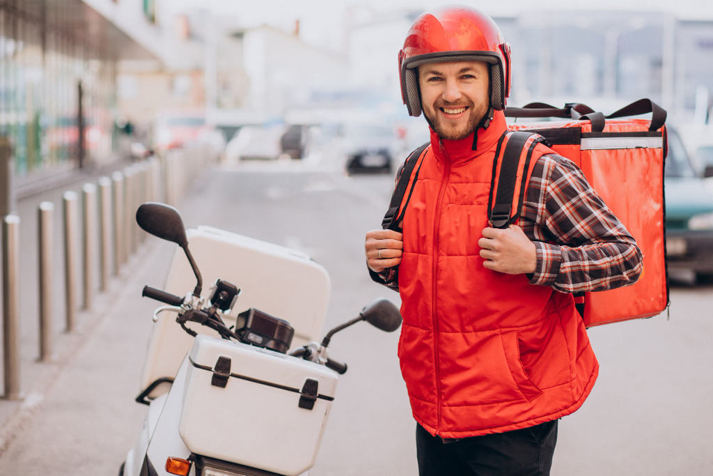 Ao lado de uma moto branca, um motoboy sorrindo com capacete, uniforme laranja e uma mala de entrega nas costas, simbolizando delivery para pastelaria