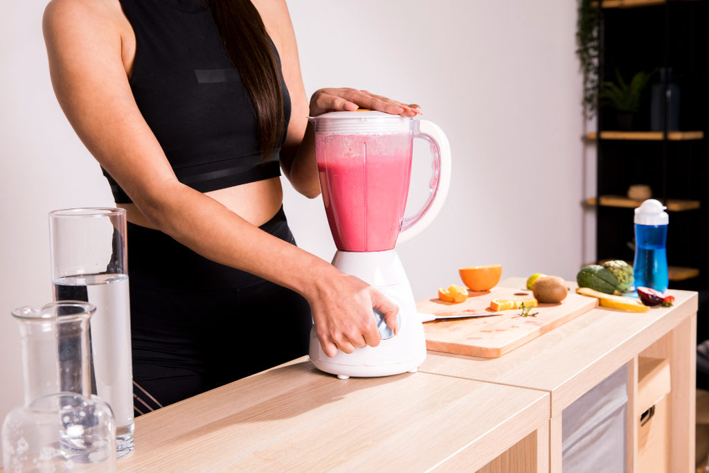 Garrafinha de água e frutas sobre a mesa, como laranjas, kiwi e abacates. Mulher com liquidificador comum sobre a mesa, batendo um líquido rosa. Simbolizando a Diferença entre liquidificador comum e liquidificador industrial