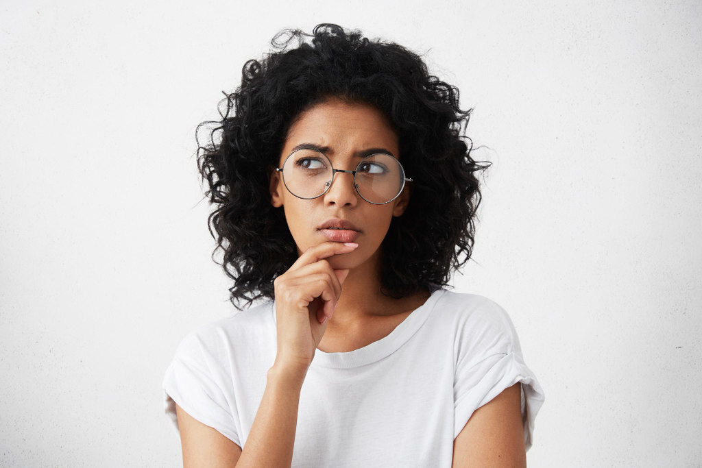 Mulher de óculos de grau com armadura redonda, mão no queixo e expressão reflexiva de curiosidade, simbolizando a dúvida sobre qual é o melhor liquidificador industrial